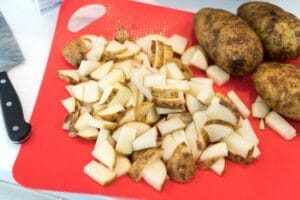 Diced potatoes on a red cutting board next to whole potatoes and a knife, solid for prepping ninja foodi steak and mashed potatoes.