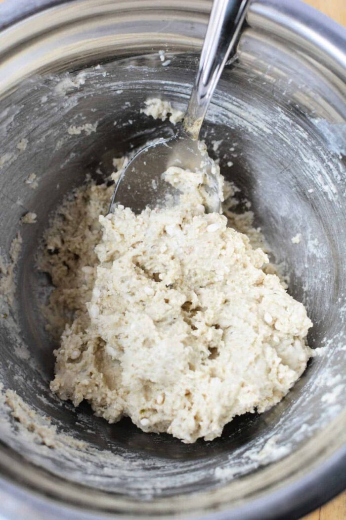 A metal bowl with a spoon mixing a pale, crumbly dough or batter mixture—solid for preparing Slow Cooker Rolls from scratch.