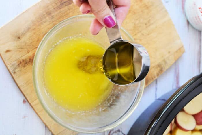 Hand pours oil from a metal cup into a bowl of melted butter on a wooden cutting board, creating the solid base for good slow cooker potatoes or other potato recipes.