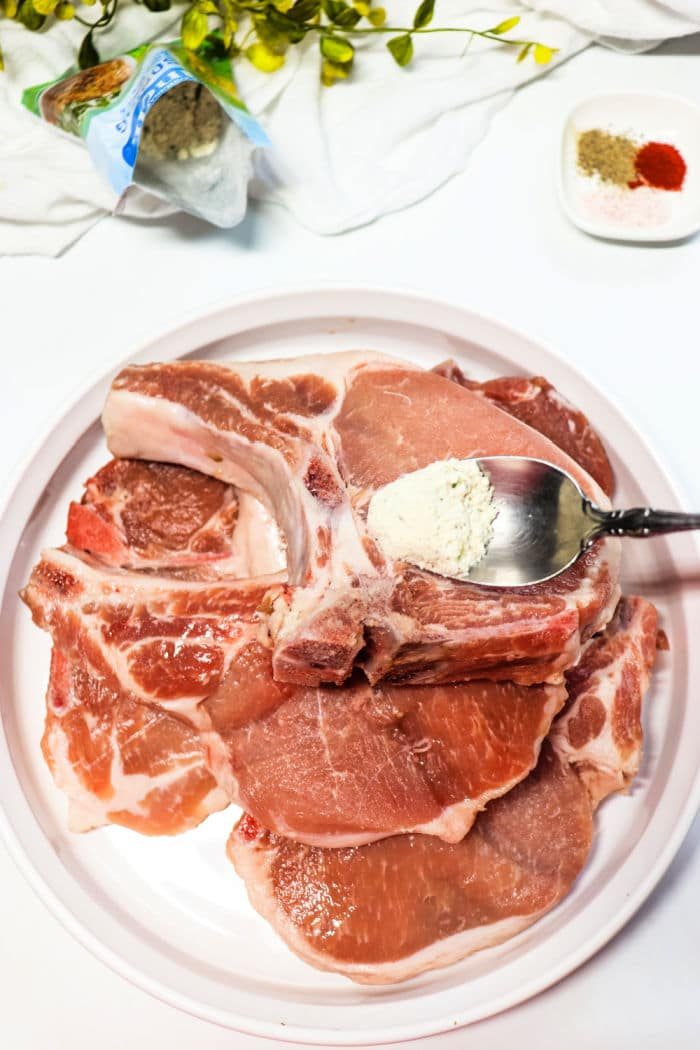 Raw frozen pork chops on a plate with a spoon adding seasoning, surrounded by spices and a leafy plant. solid for transforming into tender Slow Cooker Ranch Pork Chops that burst with flavor.
