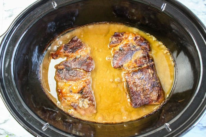 Two browned chuck roast and gravy simmers in a black slow cooker, melding rich flavors with a savory gravy.