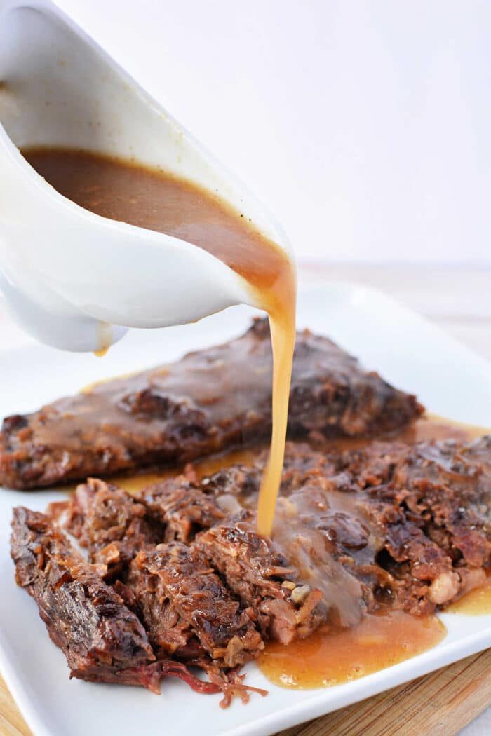 Gravy being poured from a white gravy boat over tender Slow Cooker Skirt Steak, sliced and arranged on a white plate.