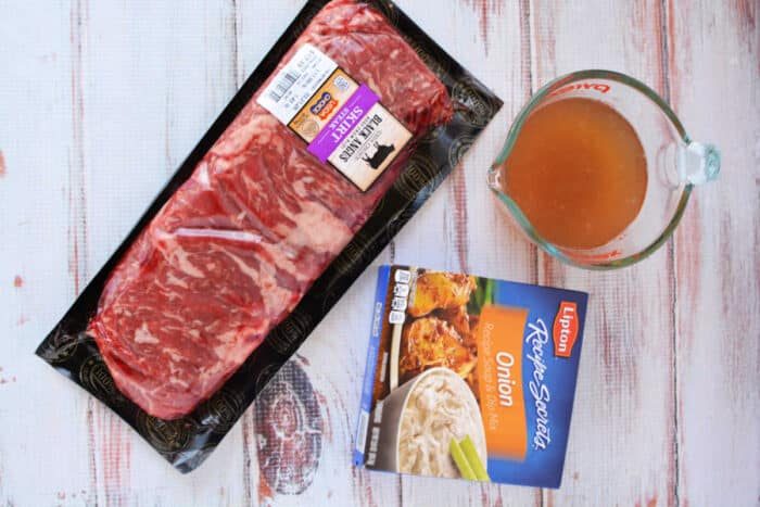 Raw beef steak in packaging, a measuring cup of broth, and a box of onion soup mix on a white wooden surface—solid ingredients for an easy Slow Cooker Skirt Steak recipe.