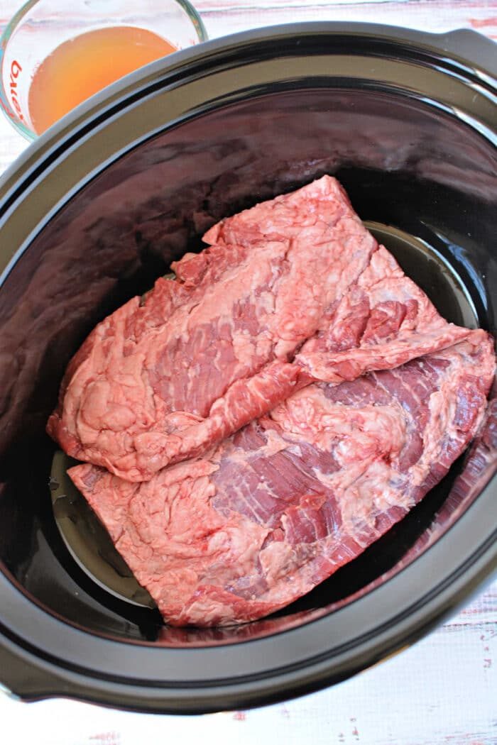 Two raw pieces of beef, ideal for slow cooker skirt steak, are placed inside a black slow cooker, with broth in a measuring cup nearby—solid for those seeking good skirt steak recipes.