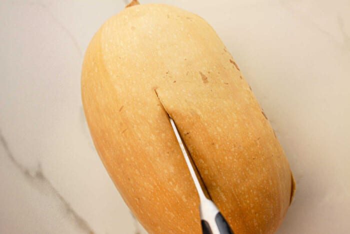 A butternut squash being sliced with a knife on a light-colored countertop, ready to transform into a good slow cooker spaghetti squash dish.