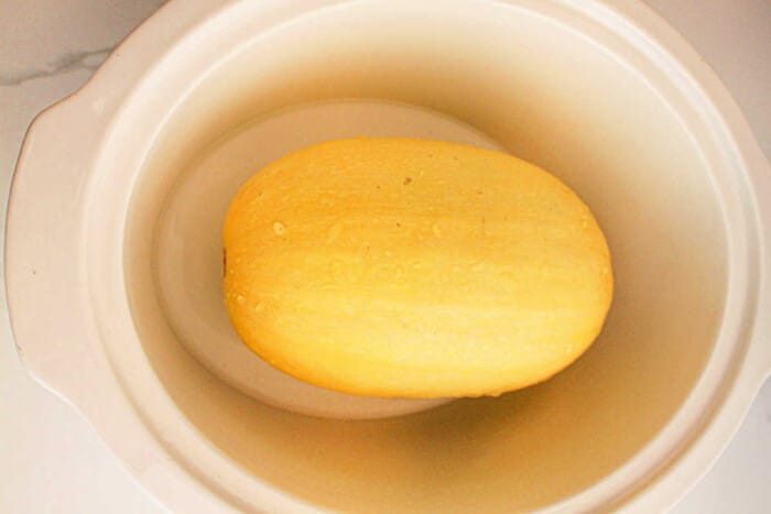 A whole slow cooker spaghetti squash sits in a white slow cooker, ready for cooking.