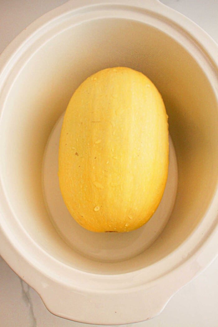 A whole slow cooker spaghetti squash nestles perfectly inside a white slow cooker, ready to transform into a delectable dish.