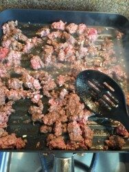 Ground beef is sizzling in a grill pan on the stovetop, solid for a mild chili recipe. A black slotted spatula rests nearby as steam rises from the browning meat. The pan's grill lines sear the beef while bits of onion add flavor, hinting at good possibilities ahead.