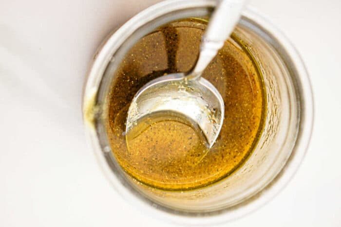 Close-up view of a glass jar filled with a golden-brown liquid, likely a sauce or dressing solid for slow cooker vegetables, with a metal spoon partially submerged in it. The liquid has some visible seasoning or spices. The background is white and minimal.