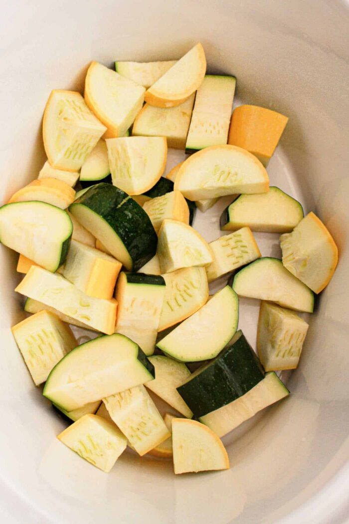 The image shows chopped pieces of zucchini and yellow squash inside a white container. The vegetables, solid for a slow cooker recipe, are cut into uniform chunks, displaying their green and yellow peels.