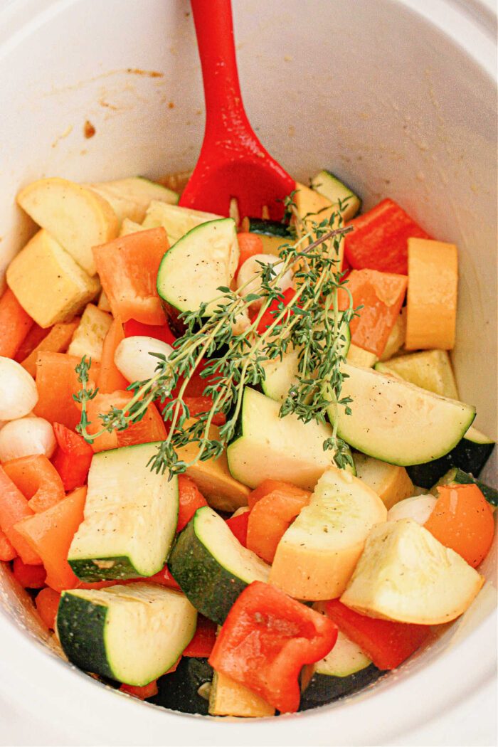 A red spoon rests in a pot filled with chopped fresh vegetables, including zucchini, bell peppers, and onions. A sprig of thyme is placed on top, adding a touch of green to the mix. The seasoned vegetables appear ready for cooking in a slow cooker.