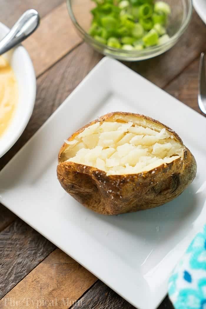 A smoked baked potato with its skin on, split open and fluffed inside, is served on a white plate. In the background, there is a small bowl of chopped green onions and a partially visible fork. The setting is on a wooden table.