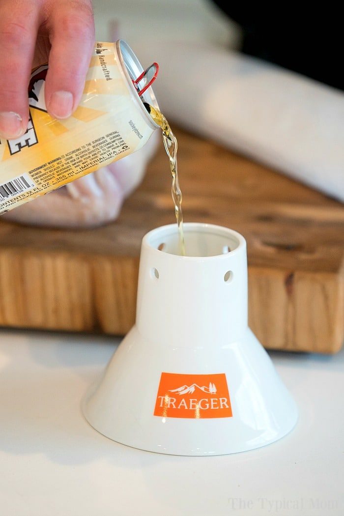 A person skillfully pours beer into a white Traeger chicken throne on a wooden countertop, preparing the solid base for a good smoked beer can chicken.