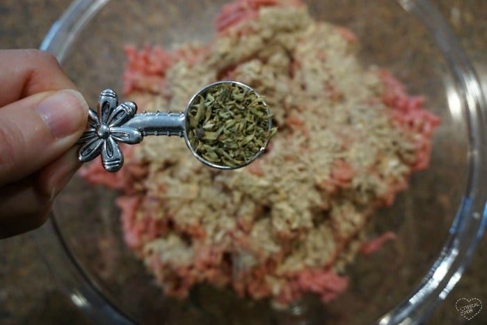 A hand holding a spoon of dried herbs over a bowl of ground meat and oats, solid for crafting a good smoked burgers recipe.