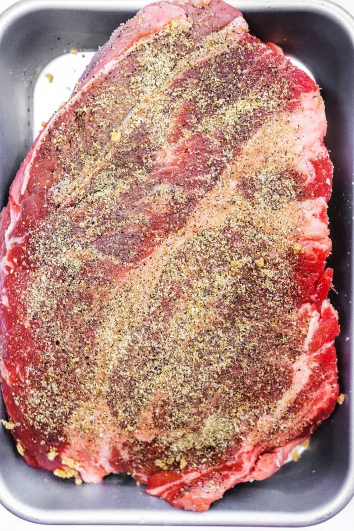 Raw seasoned beef brisket in a metal baking pan, coated with pepper and spices, ready for cooking—solid for those who love the flavors of a classic smoked chuck roast.
