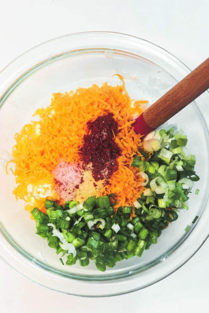 A glass bowl with grated cheese, chopped green onions, spices, and a hint of smoked jalapeño poppers, all mixed with a spoon.