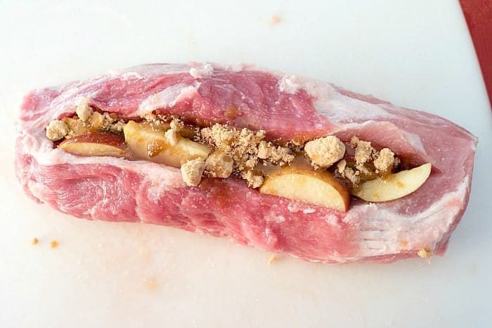 Raw pork loin, solid for a smoked pork recipe, is stuffed with apple slices and crumbled brown sugar on a white cutting board.