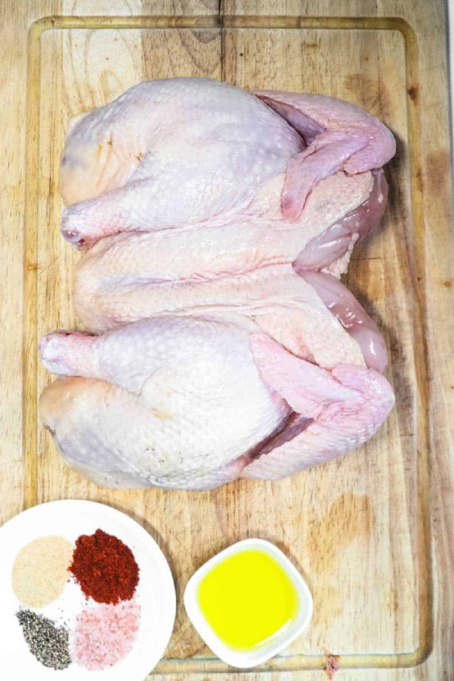 Raw smoked Spatchcock Chicken on a cutting board, with spices and olive oil in small dishes beside it.