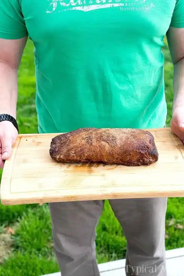 A person stands on the lush grass, holding a wooden board showcasing a perfectly smoked boneless pork loin.