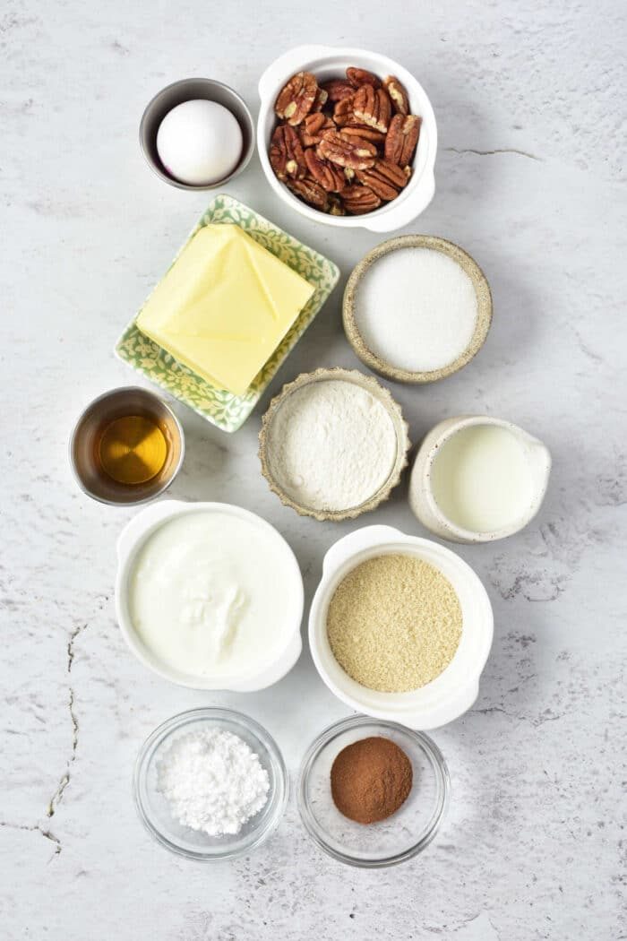 Ingredients laid out on a surface for a Sock it to us cake recipe with cake mix: egg, pecans, butter, sugar, flour, milk, vanilla, sour cream, powdered sugar, cinnamon.