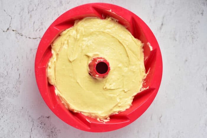 A red bundt pan filled with yellow cake batter sits invitingly on a light gray surface, ready to transform into a delightful Sock it to us cake using a convenient cake mix.