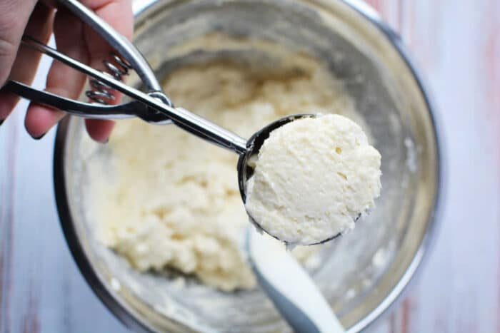 A hand holds a cookie scoop of dough over a mixing bowl with more dough and a spatula inside, solid for preparing a classic Sour Cream Biscuits recipe.