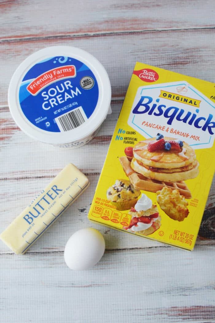Ingredients for good Sour Cream Biscuits: a box of Bisquick, tub of sour cream, stick of butter, and an egg on a wooden surface.