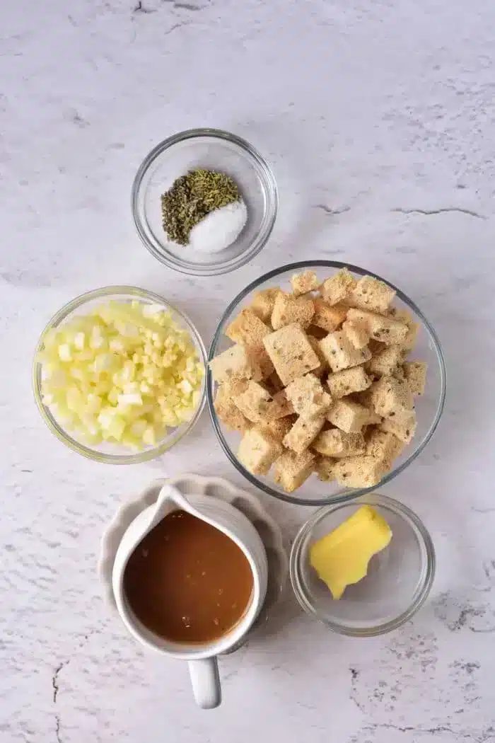 Top-down view of ingredients for a stuffing recipe, including cubed sourdough croutons, chopped onions, minced garlic, a small dish of butter, a bowl of seasoning with salt and pepper, and a gravy boat of brown liquid on a light textured surface.
