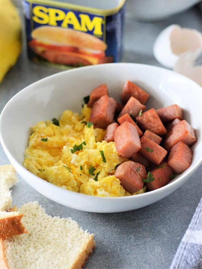 A bowl of scrambled eggs and cubed Spam, with a Spam can in the background, brings the classic flavor to life—your ideal Spam and Eggs recipe.