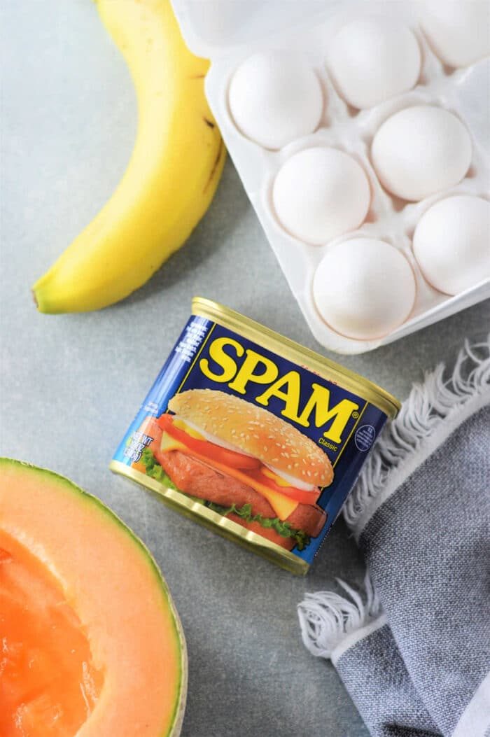 A can of Spam, a carton of eggs ready for a good Spam and Eggs recipe, a ripe banana, half a cantaloupe, and a gray cloth rest on the countertop.