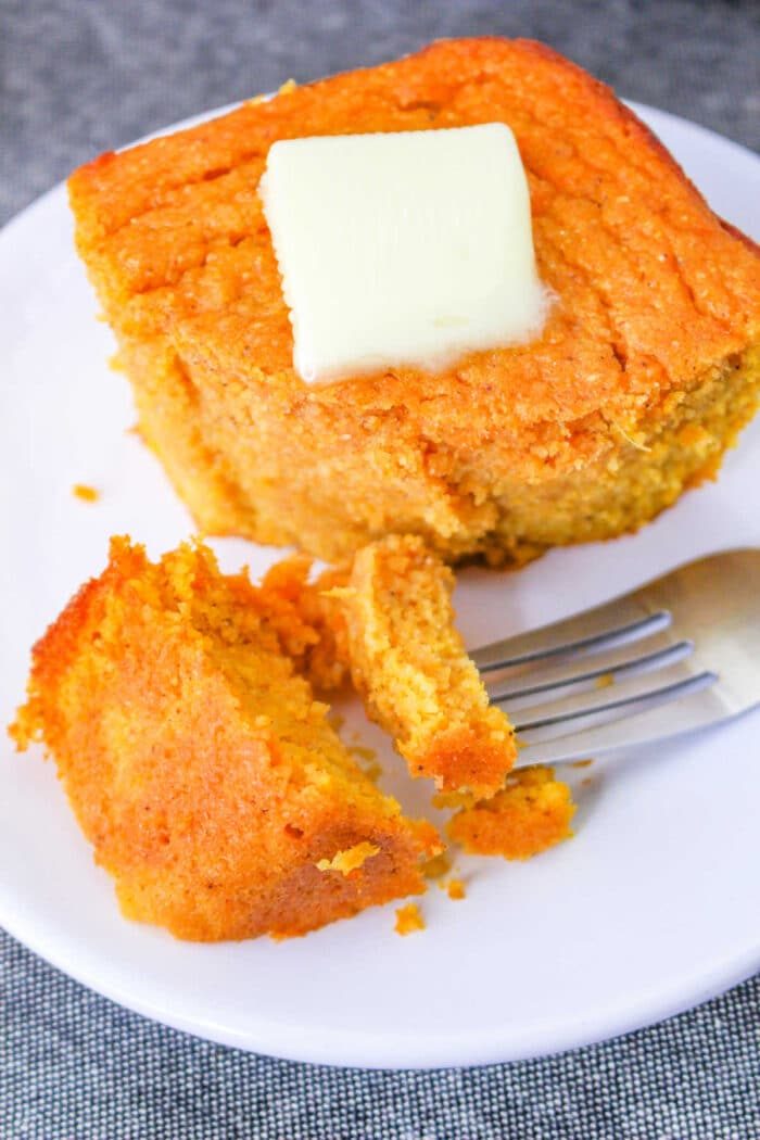 A slice of good spoonbread recipe, sits on a white plate with a pat of butter melting on top and a fork beside it.