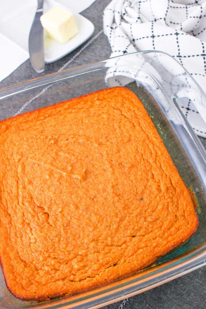 Golden-brown sweet potato corn pudding made from a classic spoonbread recipe sits in a glass dish, accompanied by a plate with butter and a checkered towel.