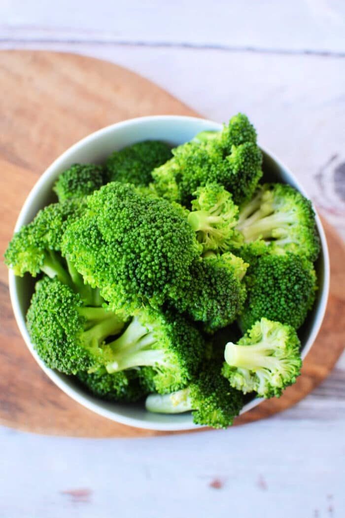 A bowl filled with fresh green broccoli florets, solid for a good steamed broccoli recipe, is placed on a wooden cutting board. The background shows a lightly textured white surface.