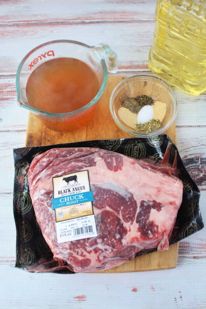 Stove Top Pot Roast ingredients: raw chuck roast, spices in a bowl, vegetable oil, and beef broth in a measuring cup all rest invitingly on the wooden surface.