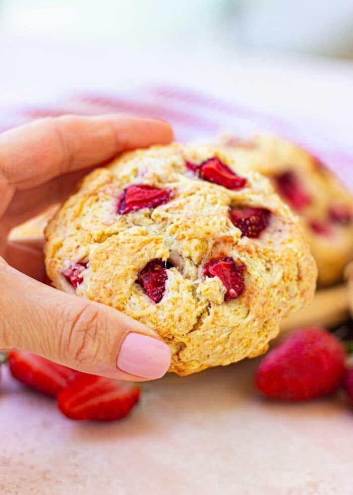 A hand holding a strawberry biscuit with visible pieces of the luscious fruit, reminiscent of a delightful strawberry biscuit, against a light background.