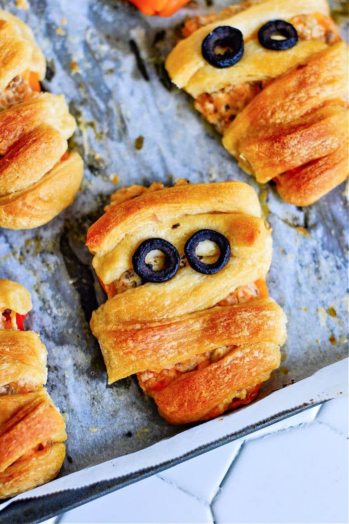 Freshly baked crescent roll mummies and stuffed mini sweet peppers rest on a parchment-lined baking sheet, the mummies shaped with olive slice eyes and wrapped dough for that classic bandage look.
