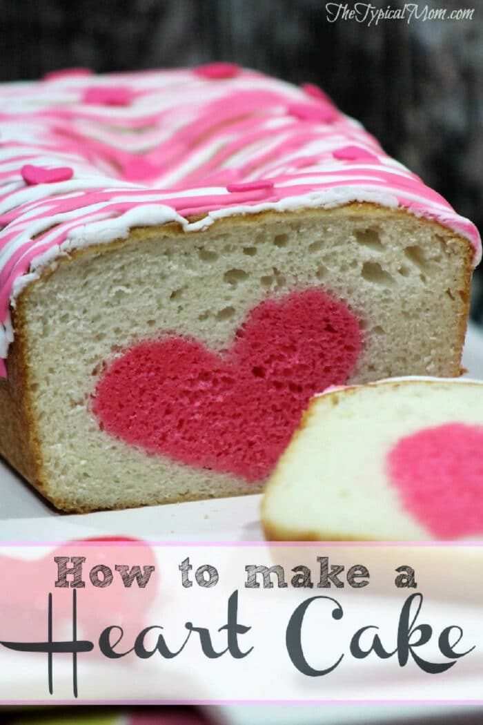 A surprise inside cake loaf with a pink heart shape in the center, topped with pink and white icing, and a sliced piece in front.