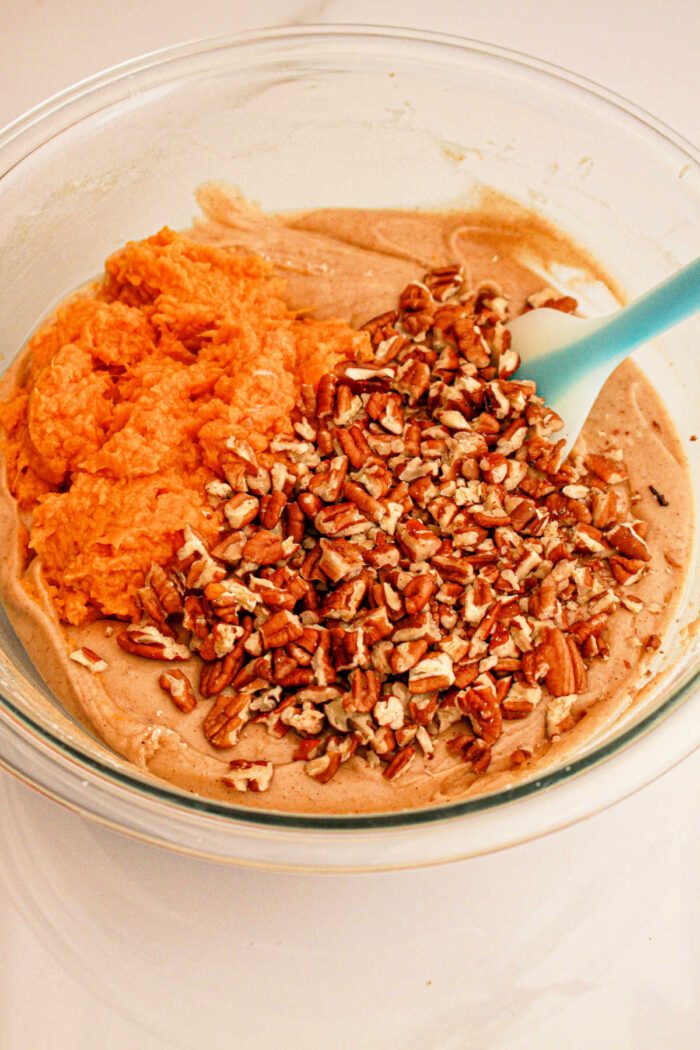 A glass bowl contains a mixture of ingredients being combined: creamy batter, orange sweet potato puree, and chopped pecans. A blue spatula is resting in the bowl, ready for mixing what will become good Sweet Potato Bread with Cake Mix.