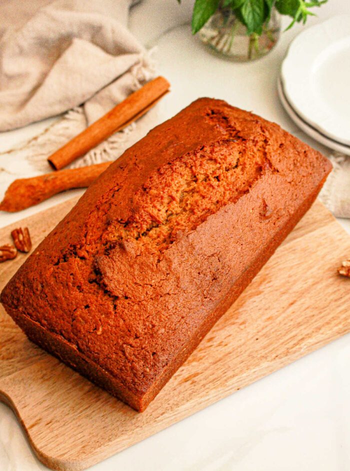 A loaf of freshly baked sweet potato bread with cake mix sits on a wooden cutting board. A cinnamon stick and some whole pecans are visible in the background, alongside a folded beige cloth and a stack of white plates.