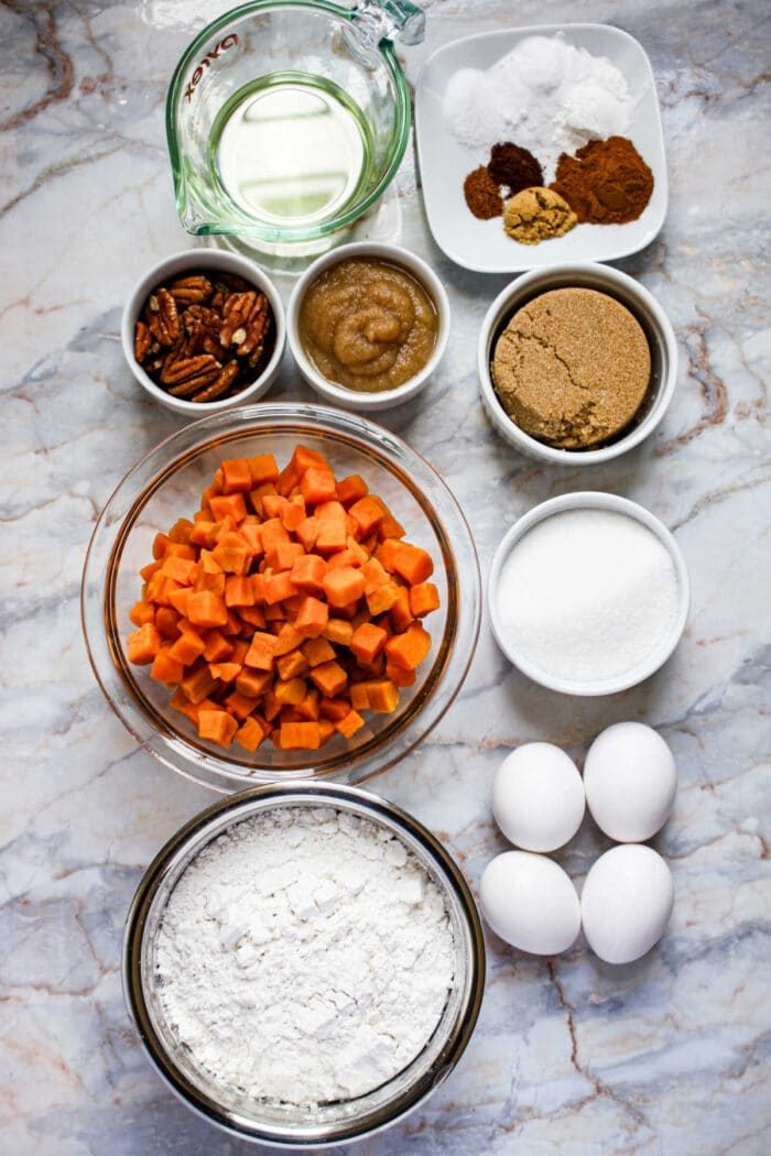 Baking ingredients on a marble surface for Sweet Potato Cake: eggs, flour, sugar, sweet potatoes, pecans, oil, applesauce, and spices.