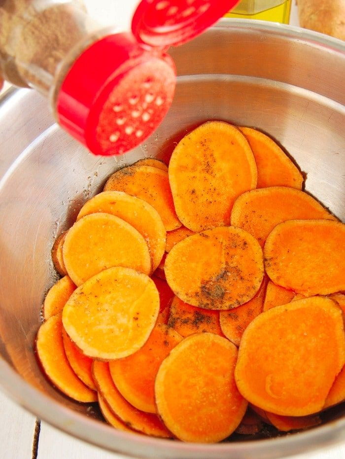 Sliced sweet potatoes in a metal bowl being sprinkled with seasoning from a red shaker, solid for making sweet potato chips in the air fryer.