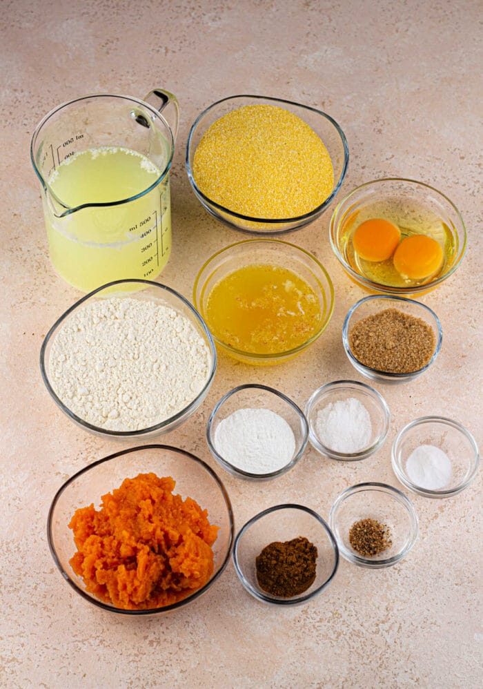 Various baking ingredients, including flour, eggs, mashed sweet potoes, cornmeal, and spices, are arranged in small bowls.