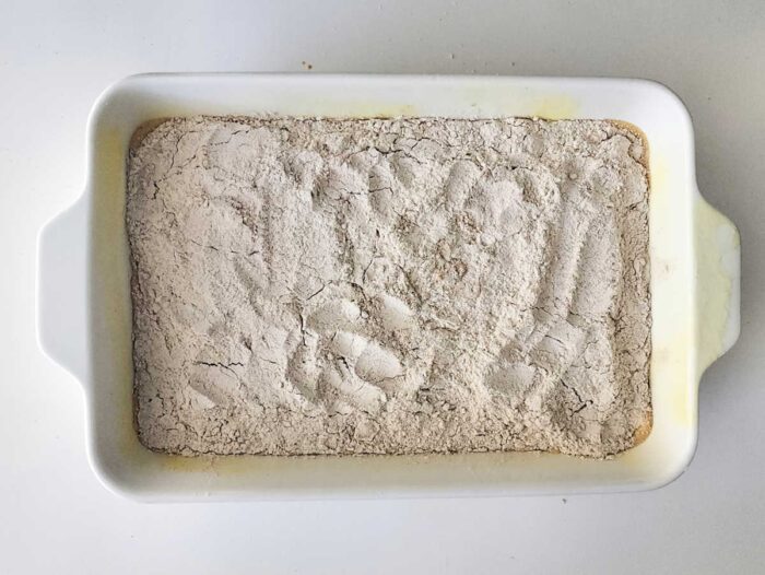 A white baking dish with a layer of flour and handprints visible on the surface, ready to become the base for a good Sweet Potato Dump Cake.