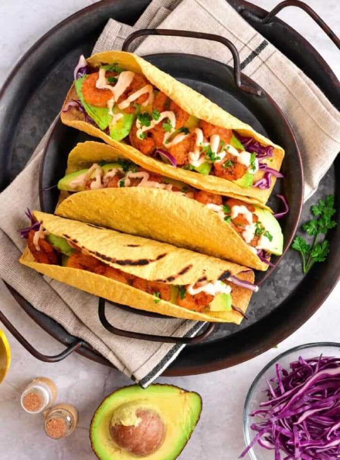 Three tacos with chicken, avocado, cabbage, and sauce sit invitingly on a plate. Beside them is a fresh avocado half and vibrant purple cabbage. These good tacos feature perfectly crispy taco shells made in an air fryer for the ultimate crunch experience.