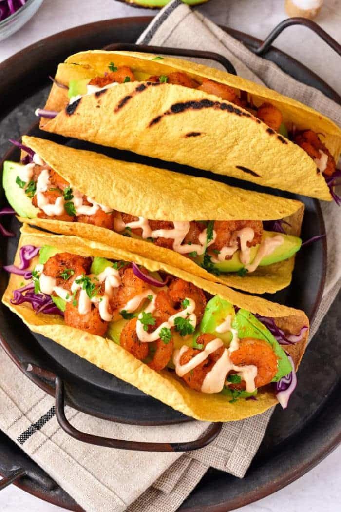 Three grilled corn tacos filled with shrimp, avocado, and cabbage, drizzled with creamy sauce on a dark plate—try making the taco shells in an air fryer for extra crispiness.