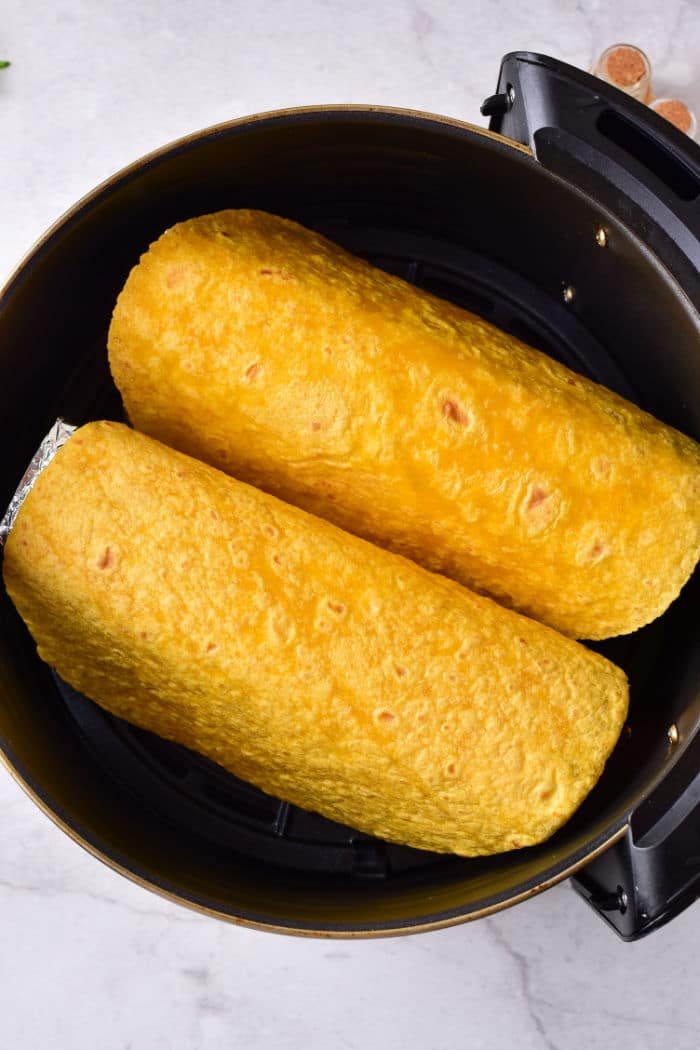 Two yellow tortilla shells wrapped around good fillings, nestled side by side in a black cooking pot, reminiscent of taco shells prepared perfectly in an air fryer.