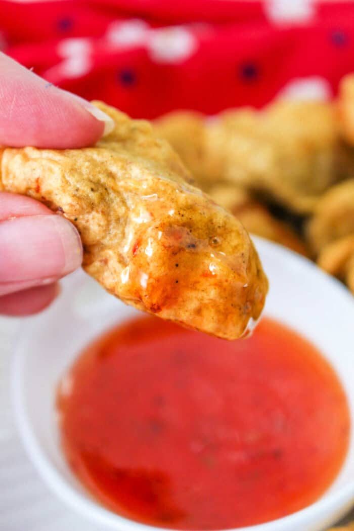 Close-up of a hand holding a golden-brown tempura chicken nuggets, dipped in a bowl of red sauce. A red cloth adds warmth to the background.