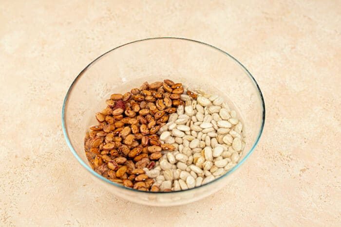 A glass bowl features uncooked tepary beans on the right, alongside white beans, all resting on a light surface.