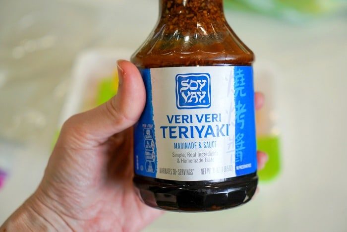 A person holds a bottle of Soy Vay Veri Veri Teriyaki marinade and sauce, solid for Traeger smoked chicken thighs. The label is blue with white text, and the bottle cap is visible at the top. The background appears to be out of focus.