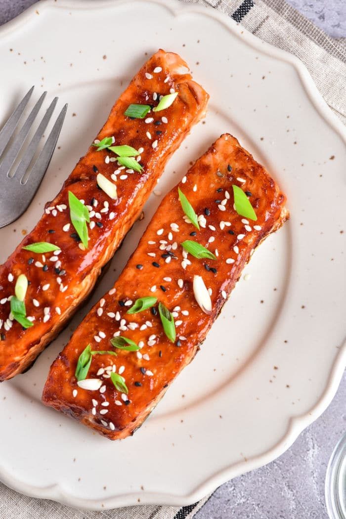 Teriyaki salmon air fried to perfection, sprinkled with sesame seeds and green onions, elegantly presented on a white plate alongside a fork.